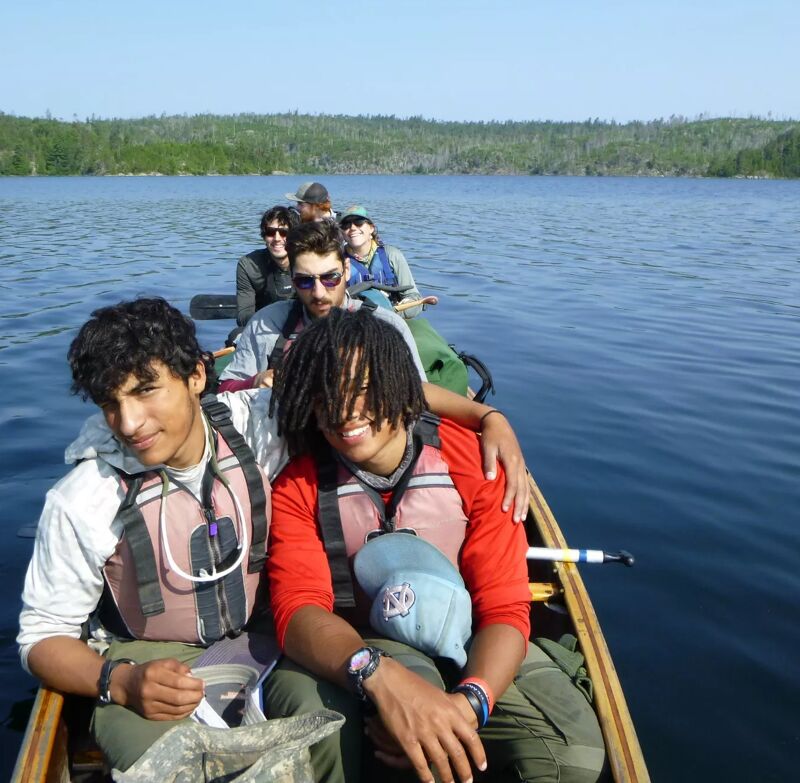 A group of people are enjoying a canoe ride on a lake. The foreground features two individuals wearing life vests, with one paddling. Behind them, other people are visible in the canoe. The background shows a treeline along the lake's edge, suggesting a natural, possibly wooded, environment. The overall scene conveys a sense of outdoor recreation and teamwork.
